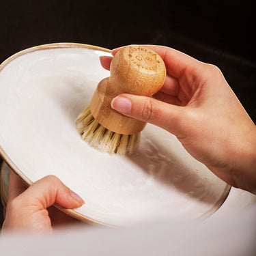 Kindred & Kin Castelbel bamboo dish brush with sisal bristles cleaning a white plate in soapy water.
