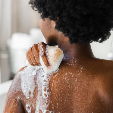 Kindred & Kin Bathorium Konjac and walnut shell exfoliating sponge creating a lather on a person's back in the shower.