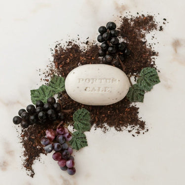 Kindred & Kin oval white exfoliating soap with grape seeds, scented with Ruby Red fragrance, surrounded by dark grapes and leaves.
