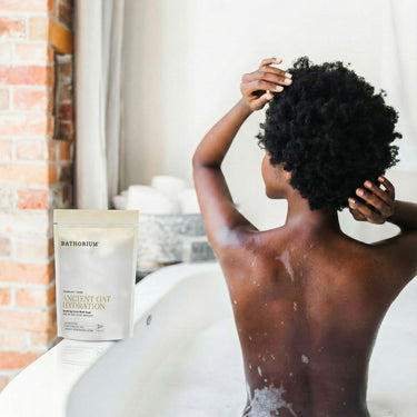 Kindred & Kin Bathorium Ancient Oat Hydration bath soak pouch next to a woman relaxing in a warm, bubbly bathtub.