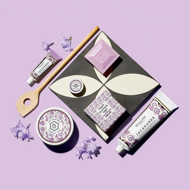Kindred & Kin Benamôr Jacarandá soap bar, body cream, and lotion set with lilac flowers on a purple background
